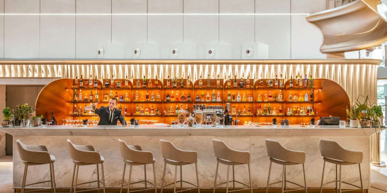 Bar du JW Marriott Cannes avec éclairage chaleureux sur bouteillerie en bois et comptoir en marbre, conception lumière Ponctuelle