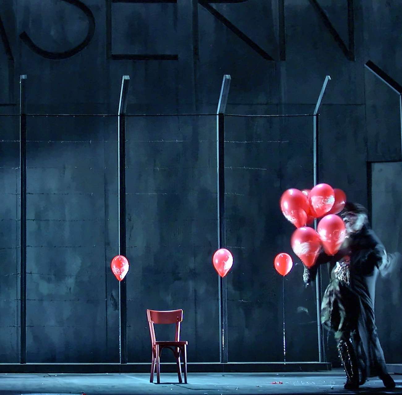 Scène de Wozzeck devant un mur de caserne gris-bleu et une clôture barbelée, ballons rouges et chaise rouge, Opéra de Nantes, conception lumière Ponctuelle