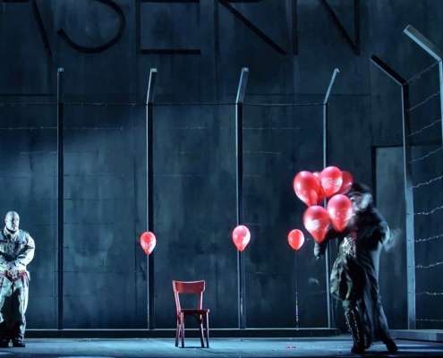 Scène de Wozzeck devant un mur de caserne gris-bleu et une clôture barbelée, ballons rouges et chaise rouge, Opéra de Nantes, conception lumière Ponctuelle