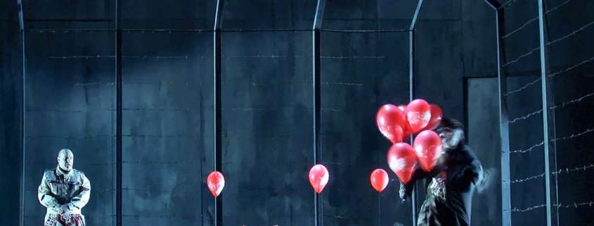 Scène de Wozzeck devant un mur de caserne gris-bleu et une clôture barbelée, ballons rouges et chaise rouge, Opéra de Nantes, conception lumière Ponctuelle