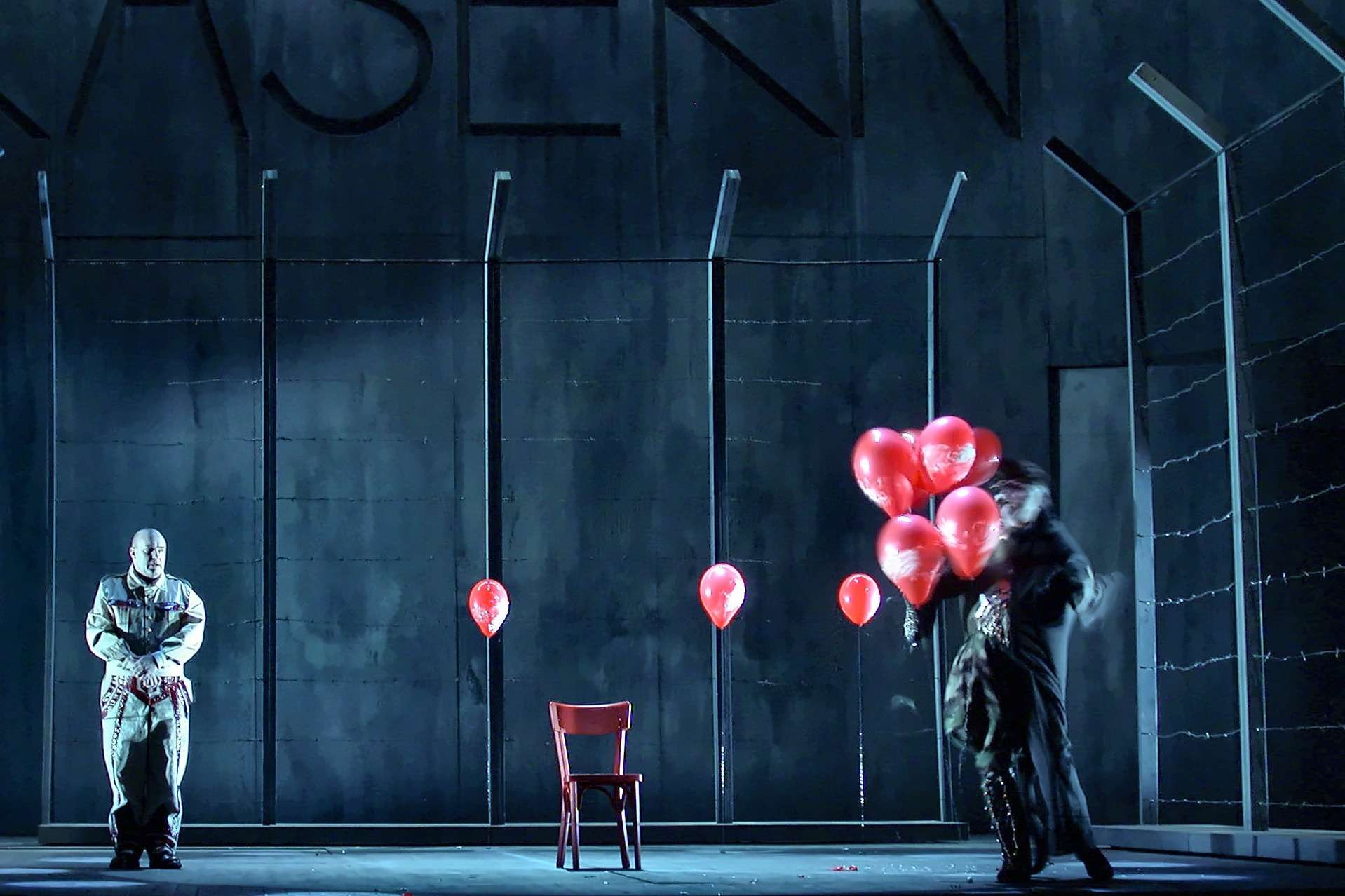 Scène de Wozzeck devant un mur de caserne gris-bleu et une clôture barbelée, ballons rouges et chaise rouge, Opéra de Nantes, conception lumière Ponctuelle